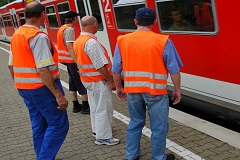 Vier Männer mit Warnwesten stehen an einem Bahngleis. Ein Zug fährt ein.